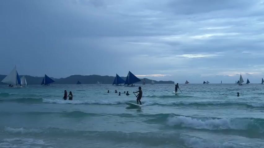 The video shows a beach scene with people swimming and paddleboarding in the ocean. Sailboats with blue and white sails are visible on the horizon. The sky is cloudy and appears to be dusk. The water is turquoise and there are waves breaking on the shore. The camera is positioned at a low angle, looking out towards the ocean.