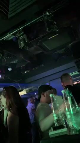 The video shows a dimly lit nightclub with a group of people standing at a bar. The ceiling is visible with lighting fixtures and air conditioning vents. Several clear bottles are lined up on the bar. People appear to be talking, but no specific interactions are discernible. The lighting is dark, mostly using green and other colored lights. The focus is on the people and the bar, with a sense of a lively night out.