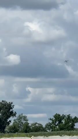 The camera looks up from a low angle at a plane in flight against a partly cloudy sky. At the bottom of the frame, a treeline is visible. As the plane flies closer, it passes over a field covered in what appears to be white plastic or netting.