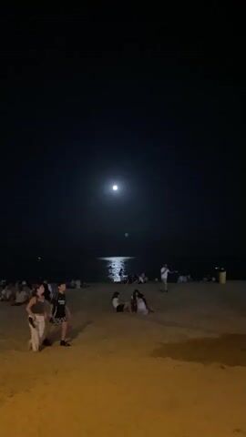 A nighttime scene unfolds on a sandy beach with a group of people gathered. In the foreground, a young woman and a boy walk across the sand. The woman is wearing a dark top and light-colored pants, and the boy is in a t-shirt and shorts. Behind them, other individuals are seated or standing on the beach. The moon is visible in the dark sky, casting a bright reflection on the calm water of the ocean in the distance. The beach is illuminated by artificial lighting, casting a warm glow on the sand.