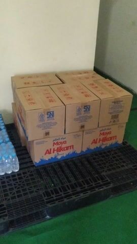 The video shows a storage area with boxes of bottled water and cardboard boxes stacked on black plastic pallets. The boxes are labeled with "Moya Al Hikam." There are multiple stacks of bottled water, each containing several rows of bottles. The cardboard boxes appear to be empty. The lighting is dim, and the walls are light-colored. The floor is covered in a green material. The video is shot from a slightly elevated, sideways angle.