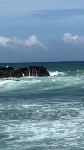 The video shows a choppy ocean with waves crashing against a cluster of dark, rocky outcrops in the center-left of the frame. The water is a deep teal color with white foam cresting the waves. The sky above is a pale blue with scattered, fluffy white clouds. The horizon line is visible in the distance, where the sea meets the sky. The overall impression is of a wild and dynamic seascape.