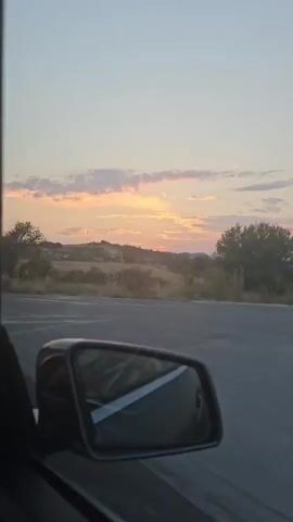 A car is driving down a road at sunset. The camera is looking out the window, and we see a side mirror reflecting the road and the sky. The sky is a pale pink and orange, with some clouds. The landscape outside the window is dry and hilly, with some trees and bushes. The road appears to be asphalt.