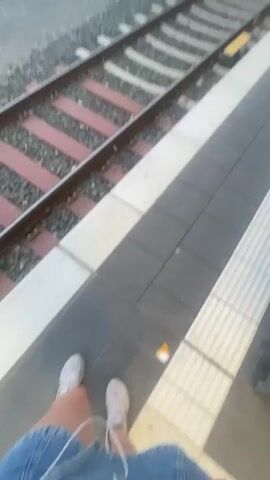 The video captures a view of a train station from a platform. In the foreground, there are train tracks with red and gray sleepers. To the right, the edge of the platform is visible, marked with black and white tiles. In the background, across another set of tracks, is a covered platform with a train stopped or moving slowly. The train is green and white. The sky is overcast and the lighting suggests either early morning or late evening. There are no people clearly visible in the video. The overall scene depicts a typical train station environment.