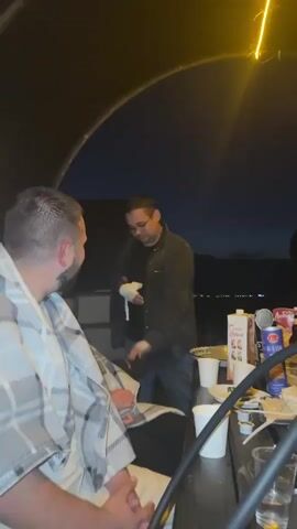 Two men are seated at a table outdoors at night. The man in the foreground is on the left, facing away from the camera and towards the other man. He has a beard and is wearing a grey and white plaid blanket over his shoulders. The second man is in the center, facing the first man. He has short dark hair and is wearing glasses, a black t-shirt, and a dark jacket. His right hand is wrapped in a white bandage. He is holding a white object in his bandaged hand and appears to be speaking. There is a table between them with several items, including cups, drinks, and what looks like food. The scene is lit by a yellow overhead light.