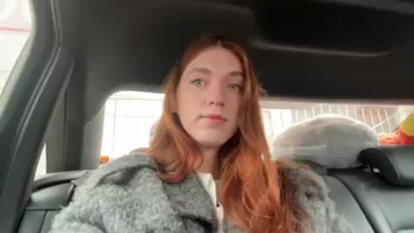 The video shows a woman with long red hair sitting in the back seat of a car. She is wearing a gray, textured coat and a white shirt. She looks directly at the camera with a neutral expression, then briefly touches her hair. Behind her, on the back seat, are a gray pillow wrapped in a net-like material, and a colorful stuffed animal. The car interior appears dark and the view through the car window is blurred.