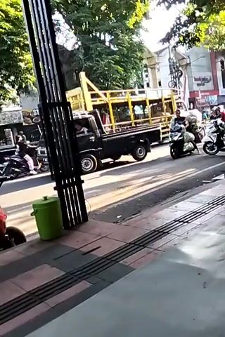 A busy street scene captured from a sidewalk. In the foreground, a black metal fence-like structure stands vertically. To the left, a green trash can is visible. The street is paved, with distinct tactile paving on the sidewalk. Vehicles are in motion on the street: a black pickup truck is seen in the background, and several motorcycles are visible. One motorcycle is carrying a significant amount of white cargo wrapped in plastic. Another motorcycle has two riders, and a third motorcycle is ridden by a single person. A yellow truck with a large cargo bed is also present in the background. Trees line the street, providing shade, and buildings are visible on the opposite side of the road. The lighting suggests it is daytime, possibly late afternoon, given the long shadows cast by the vehicles and the fence.