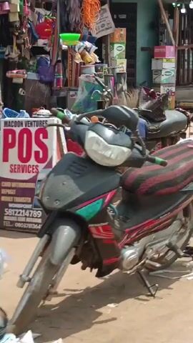 The video shows a street scene in what appears to be a busy market or commercial area, likely in an African country. Initially, two people are seen next to a motorcycle; a woman is steadying a box being placed on it by a man. Various other vehicles including a red car, auto rickshaws and a black SUV pass by in the background, suggesting the presence of active transportation. The surroundings feature modest shops and buildings. The overall impression suggests a lower-income setting with people engaging in commerce and daily activities.