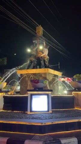 A night time video shows a fountain with a statue of three people lifting a large object. The fountain has water spraying from multiple jets. The statue is illuminated by lights, as is the fountain itself, with decorative yellow lights outlining the different levels of the fountain.
