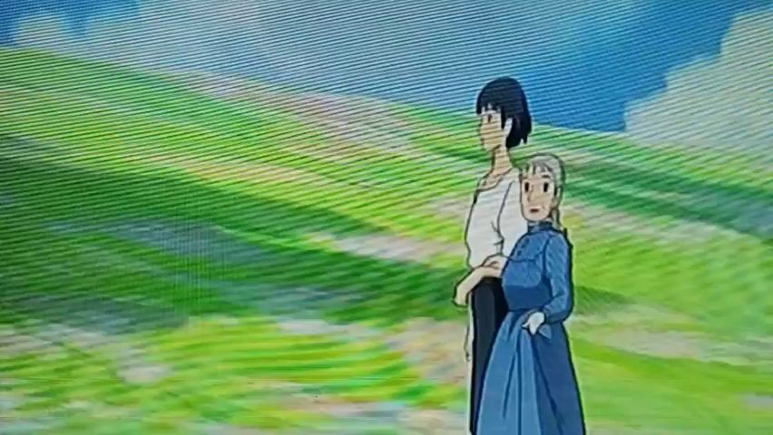 Two animated characters, a taller figure with dark hair and a lighter-skinned figure with gray hair wearing a blue dress, walk in a grassy landscape under a blue sky with white clouds. The taller figure has their arm around the waist of the other figure. The scene appears to be from an animated film. The video is grainy and appears to be a recording of a screen. Text appears at the top of the screen.