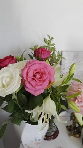 The video pans across a table displaying a collection of floral arrangements and decorative items. On the left, a white vase holds a bouquet featuring large pink roses and white lilies with green leaves. Further to the right, a lighter pink rose sits in a black tiered vase. Behind these, a square artwork with a light gray background and a white, branch-like pattern is visible. To the right of that, another bouquet is presented in a white vase decorated with faint floral imagery. This arrangement includes several yellow roses, deep red roses, and yellow lilies, all interspersed with green foliage. In the foreground, part of a dark object, possibly another vase, is visible on the far right. The surface the items are placed on appears to be white. The background is a plain white wall.
