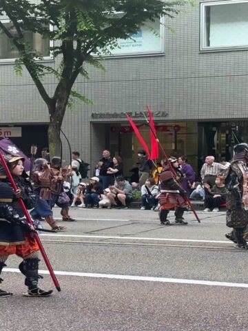 A low-angle, full shot captures a street parade with two individuals dressed as samurai warriors, facing right in the foreground, holding tall red flags on long poles with a white triangular tip. They wear traditional samurai armor, including helmets, dark blue and black chest plates, and orange hakama pants. The first samurai, positioned on the left, has a dark-colored umbrella with a white cherry blossom design tied to their back. In the background, a crowd of spectators watches from the sidewalk. A multi-story grey building with multiple windows and a sign reading "TRUST BANK" is visible behind the crowd.