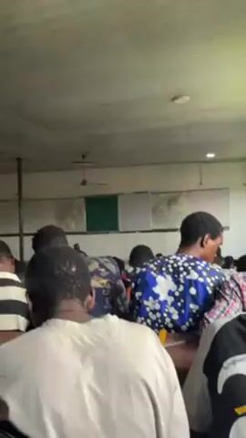 The video shows a classroom scene from the perspective of a student sitting in the middle. In the front, a man stands behind a desk, seemingly lecturing. He is wearing a colorful traditional African hat and a patterned shirt. The camera tilts up to reveal a green chalkboard with mathematical equations and notes written on it, flanked by two whiteboards also filled with writing. The classroom is filled with students, most of whom are facing the front. The room has a low ceiling with a ceiling fan. Some students are wearing patterned shirts. The overall setting is an academic environment.