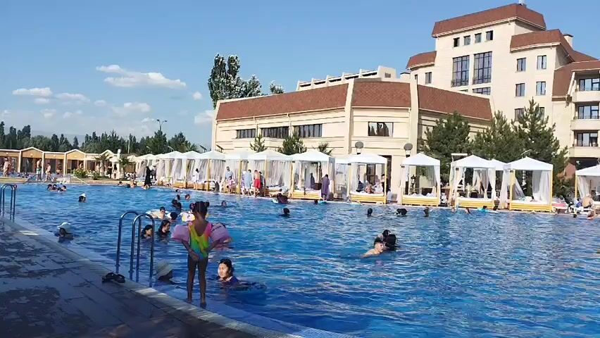 The video shows a crowded outdoor swimming pool on a sunny day. The pool is large and blue, with many people swimming and relaxing in it. There are lounge chairs and umbrellas around the pool, and a bar with people sitting at it. In the background, there is a large hotel building and trees. The video appears to be a casual recording of a vacation or resort scene.