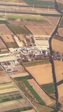 The video is an aerial shot of a rural landscape divided into agricultural fields. In the center, there is an industrial complex with several buildings, tanks, and what appears to be a water treatment facility. Wind turbines are visible in the distance, dotting the horizon across the fields. A road or canal runs along the right side of the frame. The fields exhibit different colors and textures, suggesting various crops or stages of cultivation. The lighting suggests it is daytime.