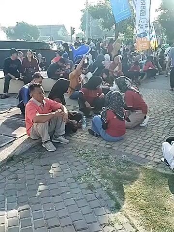 The video shows a large outdoor gathering of people, mostly adults, sitting on the ground in various groups. Many are casually dressed. Some are conversing, while others appear to be resting. There are several tents of different colors in the background, suggesting a market, fair, or organized event. The ground is a mix of paved areas and grass. The sun is shining brightly, creating lens flares in some shots. An audio track of people talking and an announcement can be heard.