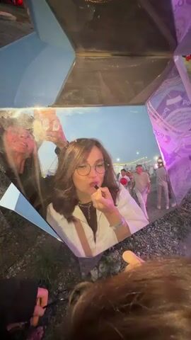A woman is applying lipstick in a faceted mirror installation. The mirror reflects a crowd of people in the background, some of whom are illuminated by stage lights. The woman is wearing glasses and a white jacket. The ground appears to be gravel or dirt.