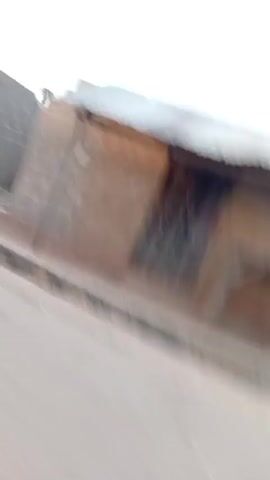 The video, apparently taken while moving, displays a view of several small, adjacent buildings. The buildings appear to be constructed of stone or brick, with corrugated metal roofing. The exteriors are in various states of disrepair and faded paint. Some have partially open doorways. A few objects can be seen inside the buildings, including what appears to be a table and some plastic crates. The road in front of the buildings is paved, but in rough condition, with gutters running alongside. The overall environment looks to be a somewhat impoverished or neglected area. The camera is moving quickly, making the details blurry and difficult to discern.