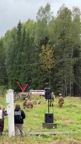 A group of people in historical military uniforms are gathered in a grassy field with trees in the background. Some individuals are positioned near a large artillery piece, while others stand or sit around the area. A speaker system is set up in the foreground. The overall scene suggests a reenactment or historical demonstration. The weather appears overcast.
