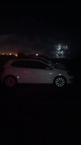 A white car is parked in a dark, outdoor environment. There are lights in the background that appear to be from a town or city. In the sky, there are several bright, circular lights that could be fireworks or other aerial illumination. The car is seen from its side, with its wheels and body clearly visible. The scene is illuminated by the ambient light from the background and the lights in the sky.