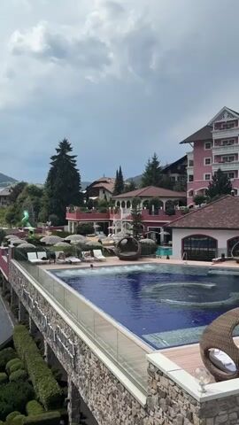 The video shows a resort with an infinity pool. People are swimming in the pool, and there are lounge chairs around it. There are also some buildings in the background, including a pink hotel. The sky is cloudy.