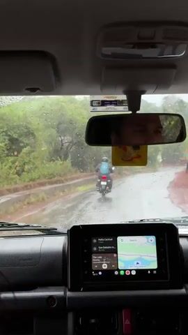 Here's a detailed description of the video:

The video is shot from the interior of a car driving on a wet road. It is raining, and raindrops are visible on the windshield. The windshield wipers are active, clearing the water.

Through the windshield, a motorcyclist is seen ahead, riding on the same road. The motorcyclist is wearing a helmet and a blue backpack. The road is lined with green trees and vegetation on either side.

The car's dashboard is visible, and the infotainment system displays navigation directions and music information. The driver's face is partially visible in the rearview mirror. A passenger's hair can be seen from the front.

The car is in motion, as evidenced by the changing scenery outside the windshield. The GPS displays the destination as Goa Dabolim and estimates the arrival time as 46 minutes.