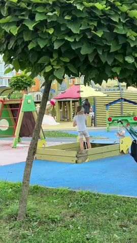 A child plays in a sandbox on a playground. The child is wearing a grey t-shirt and grey shorts. The sandbox is yellow and has a wooden border. The playground has a blue surface and green grass. There are other playground structures in the background, including a red and yellow slide and a green and yellow playhouse. A stroller is parked next to the sandbox.