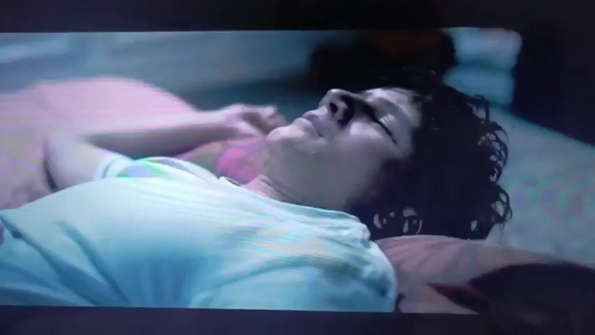 A person with curly dark hair lies on their back on a bed, wearing a light-colored t-shirt. Their eyes are closed, and their head is slightly tilted back. The person appears to be asleep or unconscious. The background is blurred but suggests a bedroom setting with bedding. The lighting is dim, with a purplish and greenish hue, creating a somber atmosphere. In a subsequent scene, a close-up shows a person's face, with dark hair and visible jewelry, appearing to interact with a baby's foot. The lighting in this shot is also low and colored, with pink and green tones.