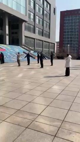 In a wide, static shot, a group of people perform what appears to be a choreographed exercise routine in a paved plaza in front of a modern, multi-story building. There are at least eight people, some wearing dark jackets or coats and one wearing a white puffer jacket. They are spread out in a loose formation, moving their arms in unison. The plaza is paved with light grey square tiles. Behind them, there is a set of wide concrete stairs leading up to the building's entrance. The stairs are decorated with colorful patterns of blue, green, yellow, and white. The building has a glass facade, and another red brick building is visible to the right. The sky is clear and the lighting suggests it's daytime. The overall atmosphere appears to be calm and orderly.