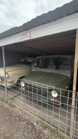 A video of vintage cars parked under a covered shelter. The shelter has a corrugated metal roof and is supported by metal poles. A chain-link fence separates the viewer from the cars. In the foreground, a green, military-style vehicle is visible. Behind it, a beige car is parked. To the right of the green vehicle, a shiny green sedan is parked. A sign above the green sedan reads "M-20 Pobeda 1956 g.v. Sdelano v SSSR" which translates to "M-20 Victory 1956 year of manufacture Made in USSR." The cars appear to be in good condition, suggesting they are either restored or well-maintained. The environment appears to be outdoors, with some gravel and vegetation visible on the ground. The sky is overcast.