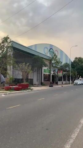 The video shows a building with the Olympic Rings logo on it, located on a street with light traffic. The building is faded in color and appears somewhat dated. There are trees lining the sidewalk in front of the building, and a few potted plants are visible. A motorcycle drives past on the street. The sky is overcast. There is a sign that says "Shop Sport". There is another sign that is unclear.