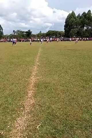 The video shows a soccer game in progress. The game is being played on a large grassy field with trees lining the back of the field and a crowd of spectators on the sidelines. There are a number of players on the field, wearing different colored uniforms. A player wearing light blue runs toward the goal. The video is taken from the sidelines and is slightly shaky.