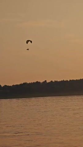 A paramotor is seen flying over a body of water near a tree-lined shore. The sky is a warm orange, suggesting it is either sunrise or sunset. The water is calm, reflecting the muted colors of the sky and land. The paramotor, consisting of a parachute-like wing and a powered backpack, appears to be the only object in motion in the frame.