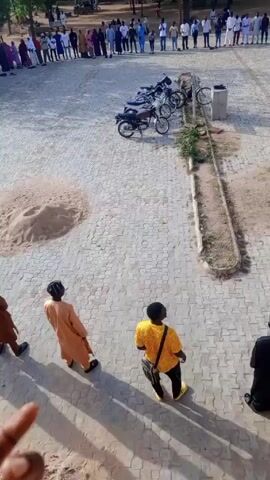 The video shows a large group of people assembled in a paved outdoor area. The group is arranged in a line, with most individuals standing. Some people are wearing traditional attire, including robes and head coverings. To the side of the group are several motorcycles parked near a concrete structure. In the foreground, three individuals are standing and observing the group. A pile of sand is visible on the paved surface. The scene is shot from an elevated position, overlooking the gathering. The video seems to capture a communal event or gathering.