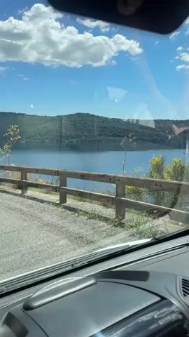 The view from inside a vehicle shows a lake and forested shoreline. The vehicle appears to be parked or moving very slowly on a road next to the lake. A wooden guardrail is visible between the road and the lake. The sky is blue with scattered white clouds.