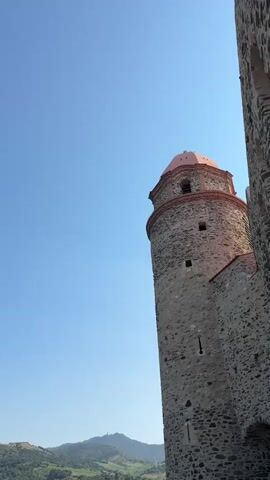 The video shows a close-up of a stone tower with a reddish dome against a clear blue sky. The tower is ancient, with rough-hewn stones forming its structure. Small, dark openings are visible at intervals along its height. To the right, another stone wall, part of the same structure, runs vertically, partially obscuring the tower. In the background, a lush green hillside dotted with buildings slopes downwards. The top of a distant mountain or hill can be seen against the horizon. The perspective is from below, looking up at the tower. The lighting suggests it is daytime, likely a sunny day. No people or specific actions are depicted.