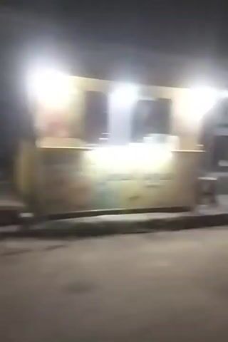 The video shows a blurry, dark street scene at night. A building with lights on is visible on the right side of the screen. There are some objects that appear to be tables or stalls in front of the building. A person on a motorcycle is seen moving quickly across the screen, also blurry. The street itself is paved, and there are some shadows and lights creating an indistinct environment. The overall impression is that of a busy, somewhat chaotic street at night, possibly a market or food stall area.