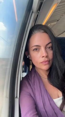 This is a close-up, first-person perspective video of a woman on a train. She is seated next to a window with a view of the sky and clouds. She has long, dark hair with some gray streaks, and she is wearing a purple scarf over a white top. She has earrings and a necklace. The train's interior is visible, including a seat behind her and overhead lighting. The camera is held at eye level, and the woman is looking directly at the camera.