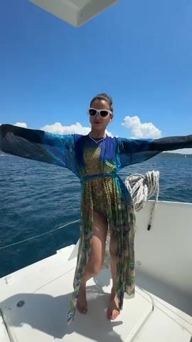 A young woman is featured in a sunlit video aboard a white boat on the sea near Bijela, Montenegro. She wears a long, sheer kaftan adorned with a vibrant peacock feather pattern over a dark long-sleeved top, and is barefoot. The sky is bright blue with scattered white clouds, and distant land or mountains are visible on the horizon.

The woman playfully poses, initially holding a dark object over her face, then revealing and adjusting white-framed sunglasses. She turns her back to the camera, spreading her arms wide and then raising them overhead, holding the sunglasses in one hand, conveying a sense of joy and relaxation in the scenic environment. A coiled white rope is visible on the boat's deck.