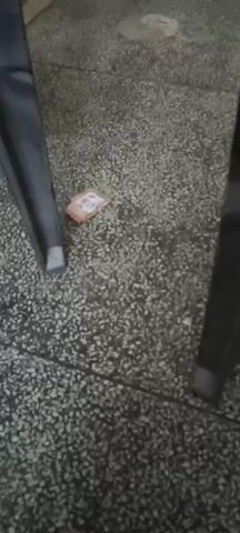 The video opens with a shot of the floor. There is some small, orange, crumpled paper on the floor, as well as what appears to be debris. Two dark chair legs are visible. The scene then shifts to a close-up view of a plastic chair. On the seat of the chair, there are two items. The item on the left is a large, light-colored bag, possibly containing food or supplements. The bag has writing and images on it. To the right of the bag is a black, cylindrical container with a black lid. This container also has writing and labels on it, including a white label with black text and a barcode. The chair and the items on it are in focus, while the background is slightly blurred. The video continues to show the chair and the items from slightly different angles, with the camera moving closer to the objects. The floor appears to be a gray, speckled surface.