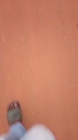A person wearing a white robe and light-colored sandals is walking on a reddish-brown surface. The camera is pointed down, showing only their feet and the ground. The video is slightly blurry, likely due to movement.