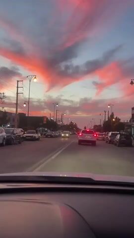 A car is driving down a city street at dusk. The sky is filled with reddish-pink clouds. The street has cars parked on both sides and other cars are driving on the street. There are street lights lining the road and power lines overhead. Buildings can be seen in the background. The video is taken from inside the moving car looking forward.