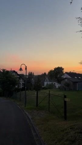 This vertical video clip captures a serene sunset over a picturesque village in Sohren, Germany. The scene unfolds along a winding asphalt road leading downhill into the heart of the village.

On the left side of the road, a well-maintained grassy embankment slopes upwards, bordered by a low stone wall. Further up, houses with traditional German architecture, featuring dark timber framing and white stucco, line the street. Some of these homes have red-tiled roofs. Mature trees provide shade and greenery. A black metal fence runs along the edge of the road, separating it from a grassy field on the right.

In the field to the right, a utilitarian shed with a wooden door stands amidst the grass. A black metal gate is visible, and further back, more houses and trees can be seen. A solitary, vintage-style lamppost with an arched arm stands prominently on the right side of the road, its dark pole contrasting with the twilight sky.

The sky is painted with the soft hues of sunset, transitioning from a pale blue at the top to warm pink and orange streaks near the horizon. This indicates the time of day is early evening. The weather appears calm and pleasant, with no visible precipitation.

Dominating the village skyline in the distance is a prominent church spire, its dark silhouette sharp against the colorful sky. Several cars are parked along the road, suggesting a residential area. The overall atmosphere conveyed is one of tranquility and peacefulness, typical of a quiet European village at dusk. No visible text is present in the video.