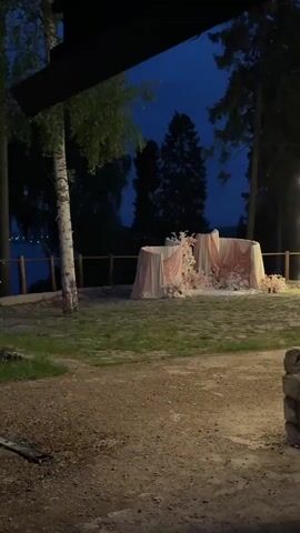 A close-up shot captures a wedding ceremony setup in an outdoor setting at dusk. The arrangement features several tall, cylindrical structures draped with white and pink fabric, forming a semi-circular backdrop. These structures are adorned with numerous white and pink floral arrangements, creating a romantic and elegant atmosphere. The ground in the foreground is covered with green grass and some scattered dirt. In the background, dark trees stand against a twilight sky, adding to the serene ambiance. The overall lighting is dim, suggesting the event is taking place in the early evening.