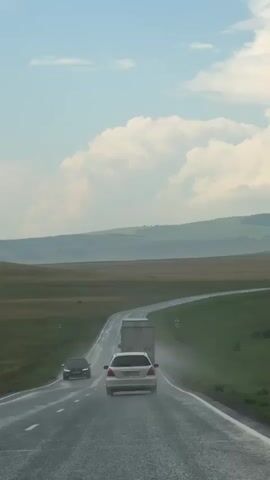 The video shows a view from inside a vehicle looking out at a highway. The road is wet, indicating recent rain, and a white car and a truck are driving ahead. The landscape is open with green fields and rolling hills under a cloudy sky.