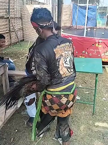 A man in a black shirt, jeans, and a black hood is being carried by a man with a painted face, a black shirt with a tiger design on the back, and a patterned sarong around his waist. The scene appears to be outside under a structure, possibly a tent or open-air building. There are other people around, some of whom appear to be watching the interaction. The carried man has his head down and seems compliant. The man carrying him is walking steadily, with some purpose, towards a corner of the structure. The environment includes rustic benches, a bamboo fence, and a tarp. The overall atmosphere seems casual and social.