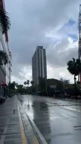 The video shows a street scene after a rainfall, indicated by the wet pavement reflecting the sky. A tall building stands in the background, with a construction site nearby where a building is under construction. A large crane is visible at the construction site. Trees line the street, including palm trees and an evergreen tree. A building with red accents is also visible on the left side of the street. The sky is cloudy.