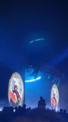 The video shows an outdoor concert stage from the perspective of the audience.  There are two large circular screens displaying silhouetted figures on either side of a central platform.  The platform is supported by a large metal structure with lighting fixtures.  Blue lighting and fog effects are prominent. The audience is visible in the foreground, with many people holding up their phones. The sky is dark, with a few visible lights. The camera angle is tilted upwards, capturing the height of the stage setup.