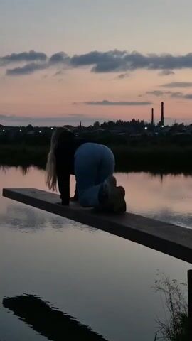A person with blonde hair wearing a dark jacket and light blue jeans is on all fours on a wooden plank that extends over a body of water. The person's back is facing the camera, and they appear to be moving forward on the plank. The sky in the background is dusky with clouds, and there are distant structures visible on the horizon. The water reflects the sky and the plank.