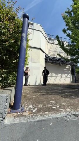 The video features two young men performing scooter tricks in an urban outdoor setting in Bron, France, under sunny skies.

The first segment shows a low-angle shot of a young man, dressed in a black t-shirt, dark pants, and a black beanie, riding a scooter. He approaches a blue metal pole and performs a bunny hop, clearing the obstacle. Another individual on a scooter is visible in the background next to green bushes. A modern, light-colored building with an angular design is in the background.

The second segment captures another young man, viewed from behind, wearing a black long-sleeved shirt with "CARR 4" printed on the back, dark pants, and blue and yellow Adidas sneakers. He rides his scooter across a paved area, crouching low over the scooter's deck. The scooter deck has "KOSTYA HOLOV" visible. The background features a distinctive modern building with geometric facades and large windows. Part of a sign on a glass door reads "de l'Université," indicating a university campus. A rolling suitcase is seen near the building, and trees dot the landscape. The sky is bright blue with visible contrails.